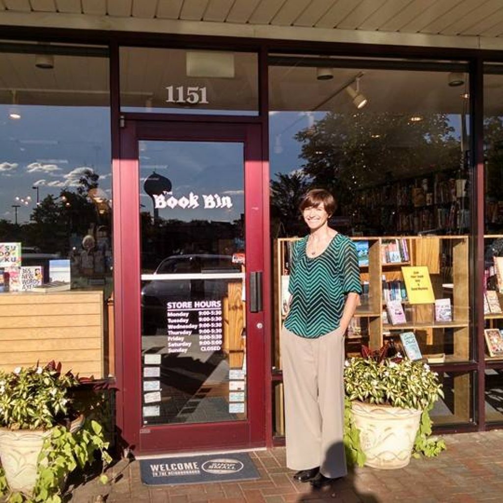 A woman smiling outside a local bookstore named "book bin" on a sunny day, with books displayed in the window and benches nearby inviting passersby to sit and enjoy the area.