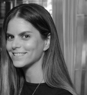 Woman with long straight hair smiling, wearing a dark top, in a black and white indoor setting.