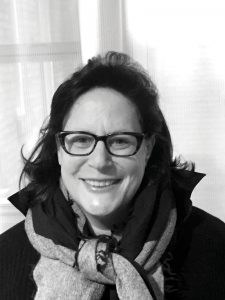 A black-and-white portrait of a smiling woman with glasses, wearing a scarf and a black top.
