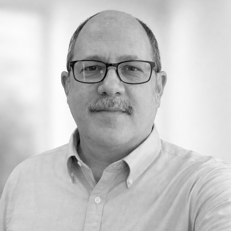A man with glasses and a mustache wearing a light button-up shirt, standing in a bright, blurred background.