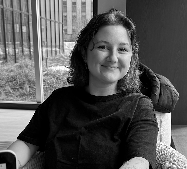 A person with wavy hair smiles while sitting indoors near large windows and wearing a dark t-shirt.