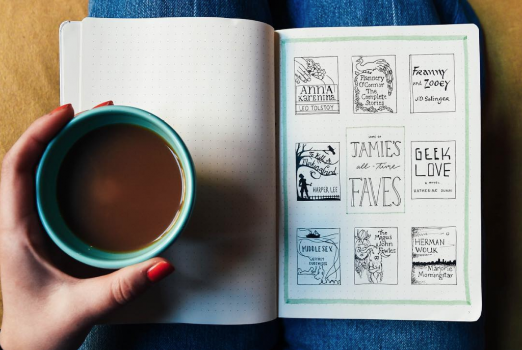 A person holding a cup of coffee while showing a journal page featuring doodles and titles of favorite books.