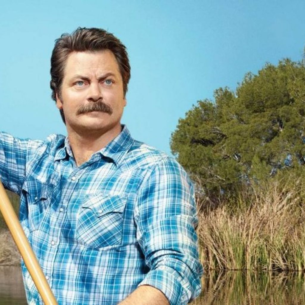 A rugged individual with a mustache, wearing a blue plaid shirt, confidently holds a paddle while standing in a red canoe, amidst a scenic backdrop of lush greenery and clear blue skies.