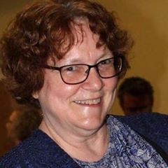 A smiling woman with curly hair and glasses, wearing a blue sweater and a patterned blouse.