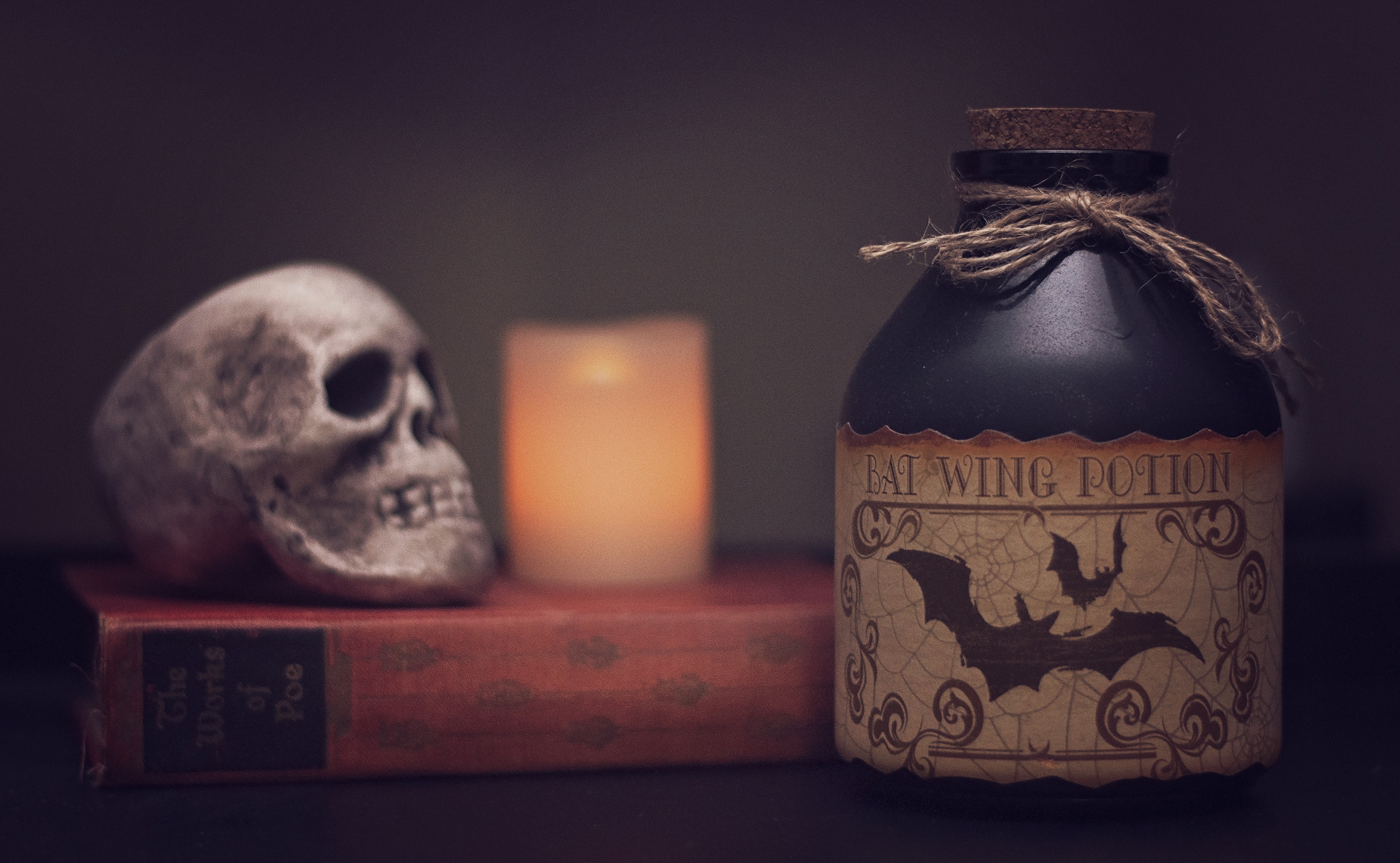 A mysterious and moody still life scene featuring a human skull resting on an old book, beside a candle, and a bottle labeled "batwing potion" with halloween-themed decorations.
