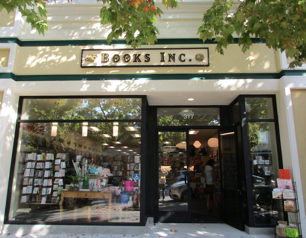 A sunny day outside the inviting entrance of books inc., with an array of books visible through the large glass windows, inviting passerby to discover a world of reading inside.