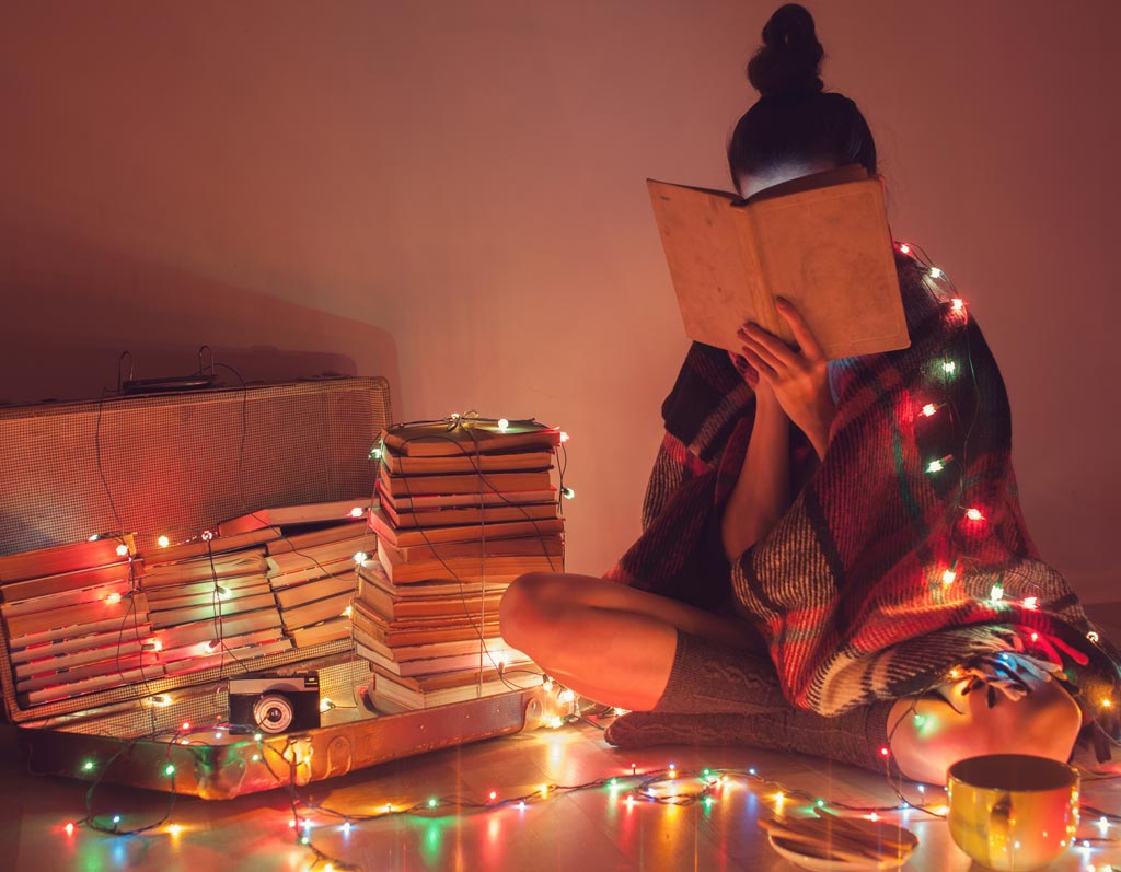 A cozy reading nook illuminated by the soft glow of fairy lights, with a person engrossed in a book amidst stacks of literature and the warm ambience of a peaceful evening.