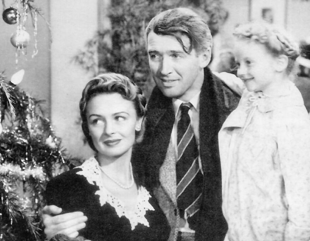 A black and white photo of a classic film scene showing two adults and a child sharing a warm, joyful moment next to a christmas tree, evoking a sense of holiday nostalgia.