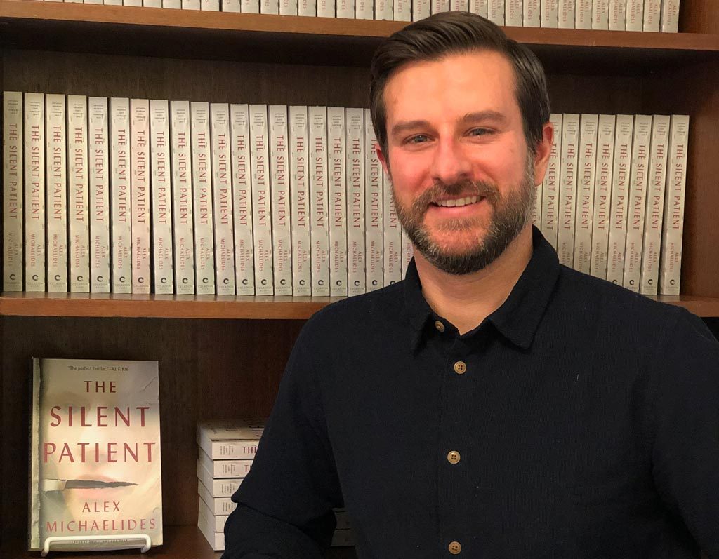 Man smiling in front of a bookshelf filled with copies of "the silent patient" by alex michaelides.
