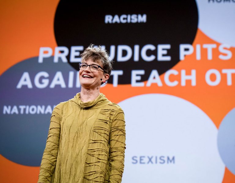 A speaker stands confidently in front of a presentation screen with the message "prejudice pits us against each other," surrounded by words like "racism," "sexism," "homophobia," and "nationalism.