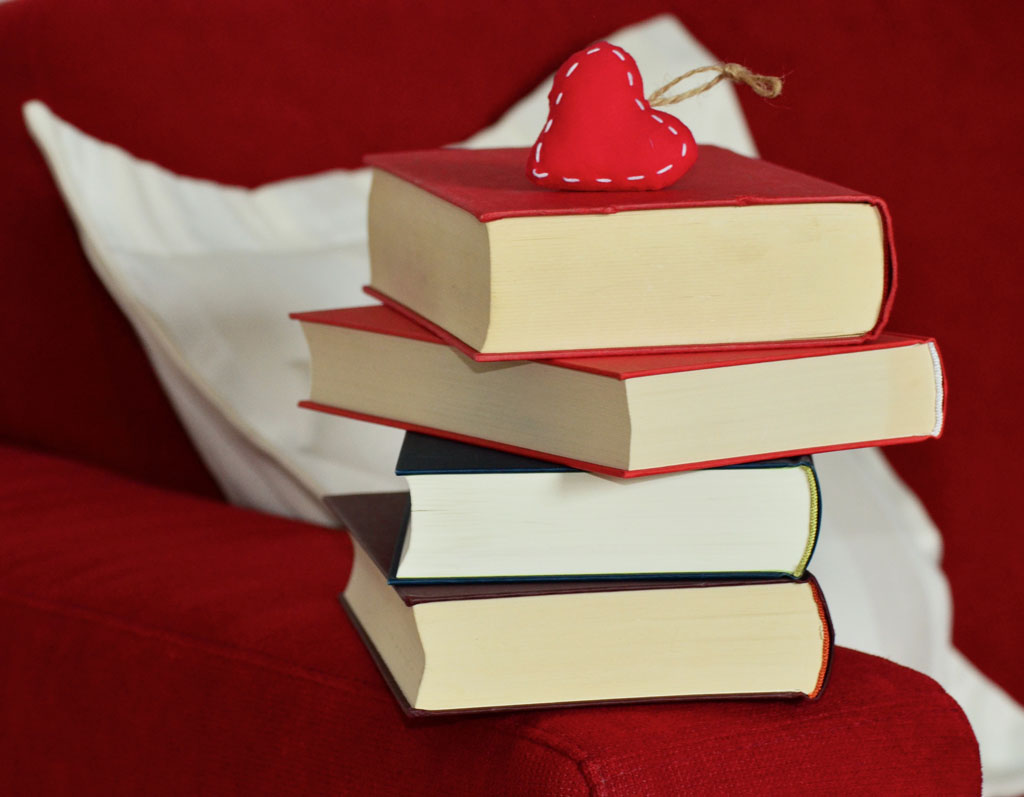 A stack of five hardcover books on a red couch with a plush heart on top, suggesting a love for reading or a cozy reading corner setup.