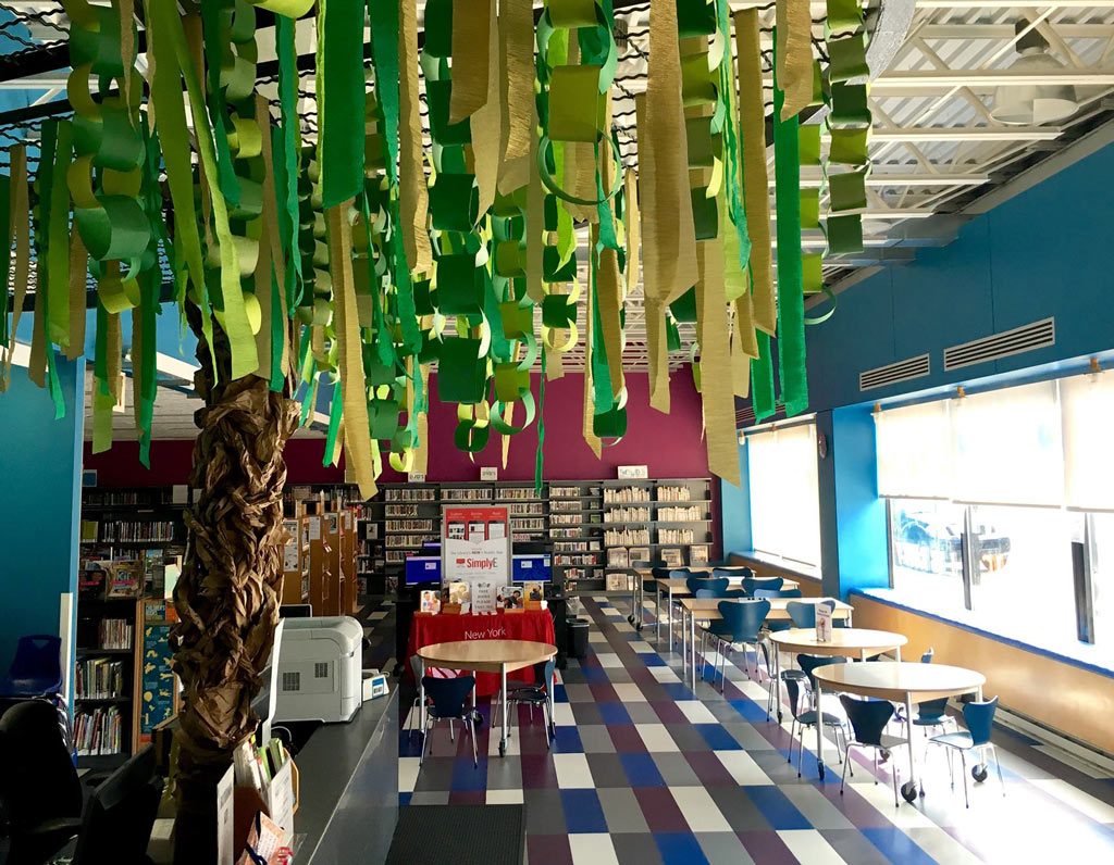A vibrant and colorful library space with hanging green decorations, possibly suggesting a theme or a special event, against a backdrop of bookshelves, tables, and chairs inviting reading and learning.