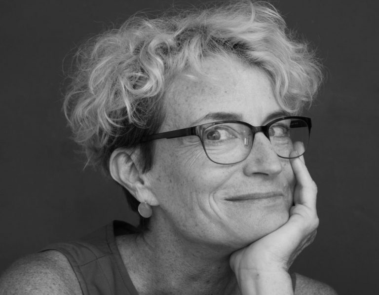 A black and white portrait of a smiling woman with short curly hair, wearing glasses, and resting her cheek on her hand.