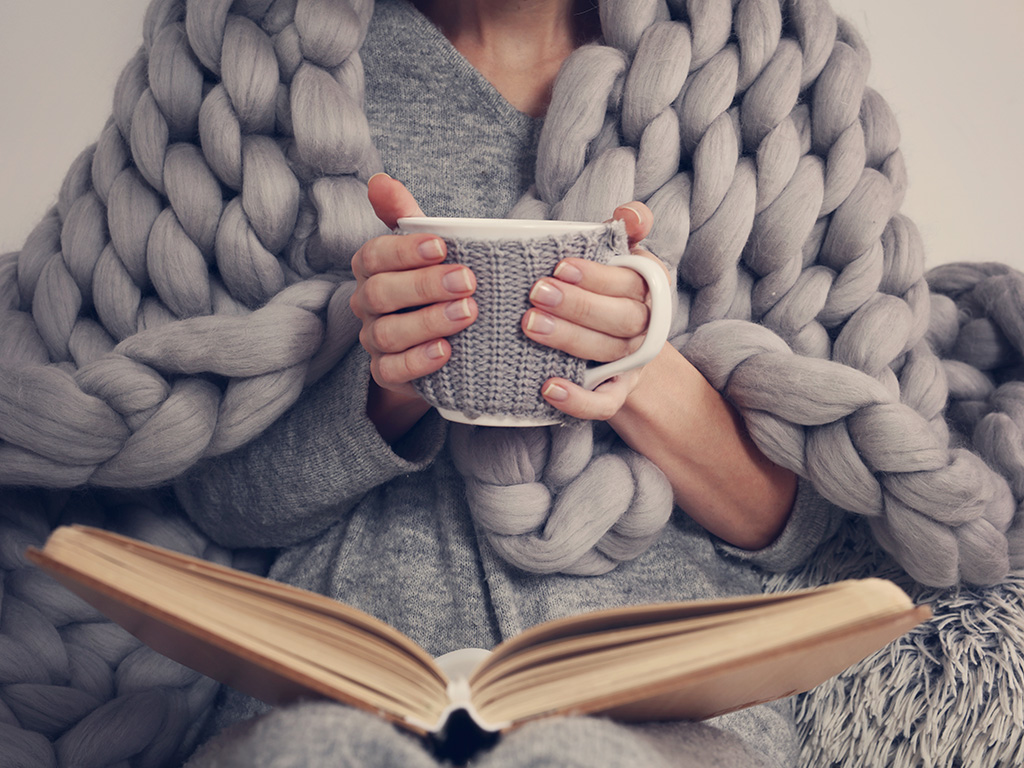 Cozying up with a warm drink and a good book, wrapped in a chunky knit blanket.