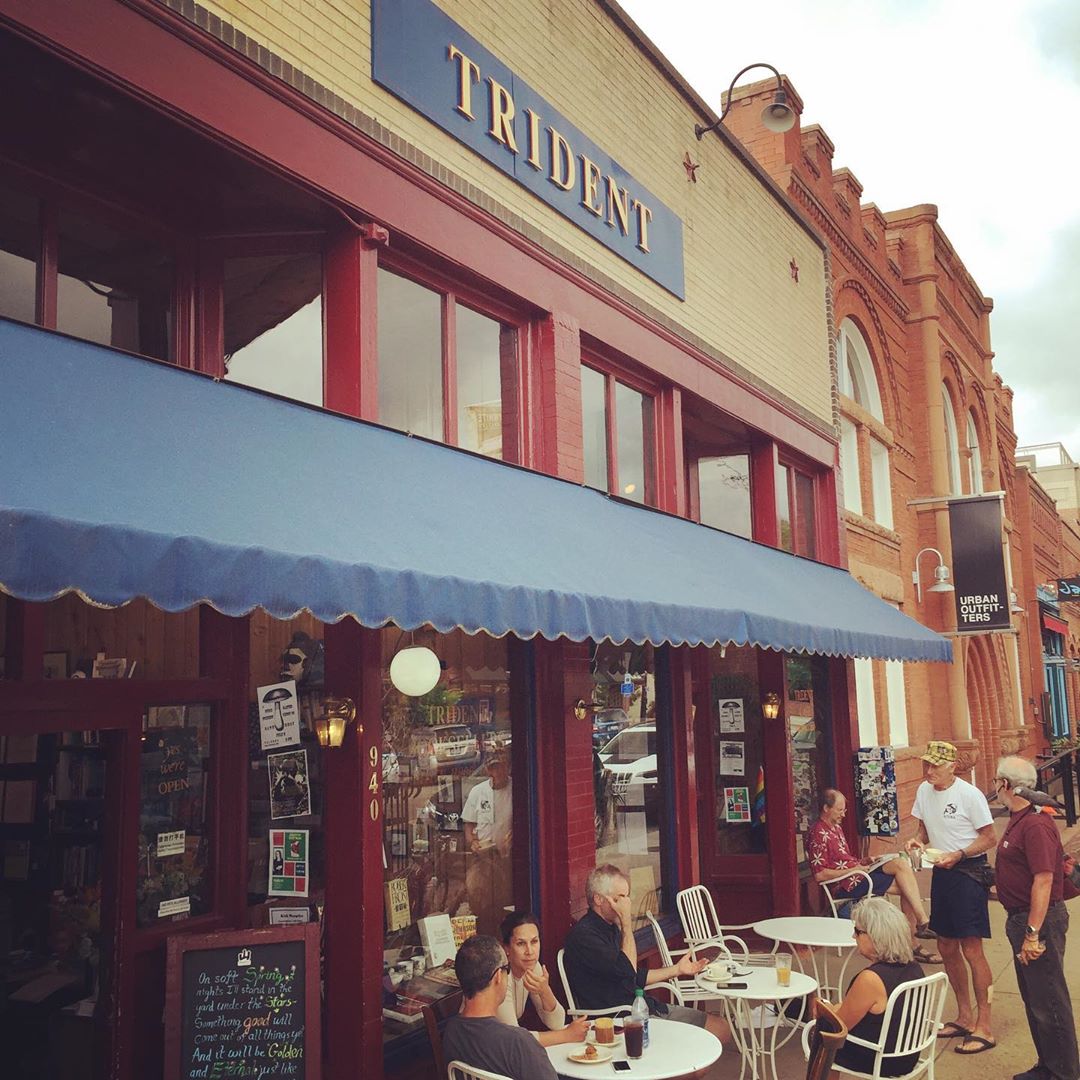 A lively street scene featuring patrons enjoying outdoor seating at the trident bookstore and cafe under a blue awning, with the warmth of a brick building façade in the background.
