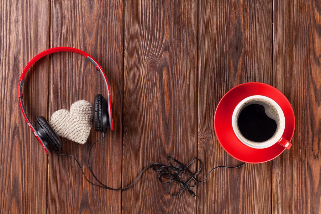 A cozy arrangement featuring a pair of headphones lovingly placed around a knitted heart next to a red cup of coffee on a wooden table.
