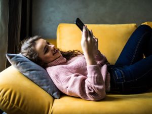Young woman reclining on a yellow sofa, smiling as she engages with her smartphone.