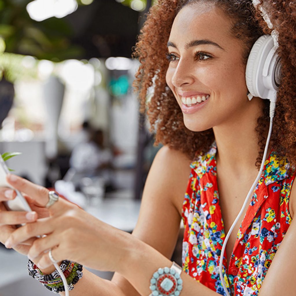 A joyful young woman wearing headphones and using her smartphone at a cheerful outdoor cafe.