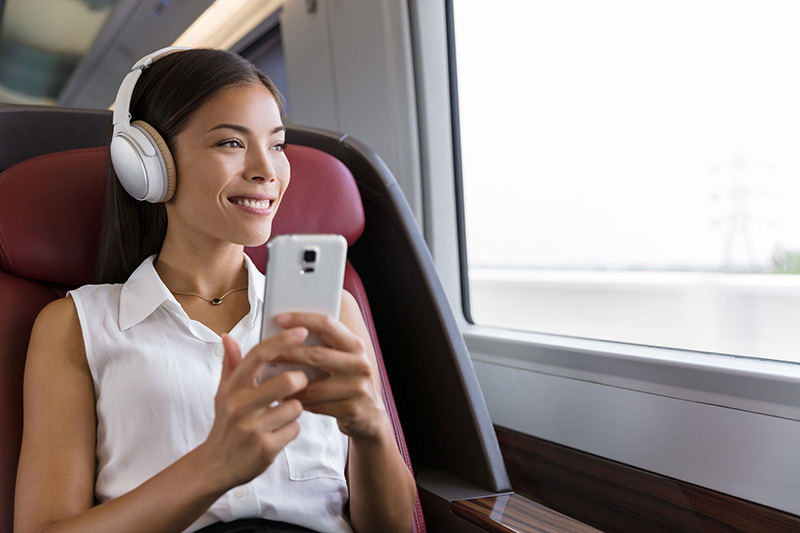 A smiling woman wearing headphones enjoys looking at her smartphone while traveling on a train.