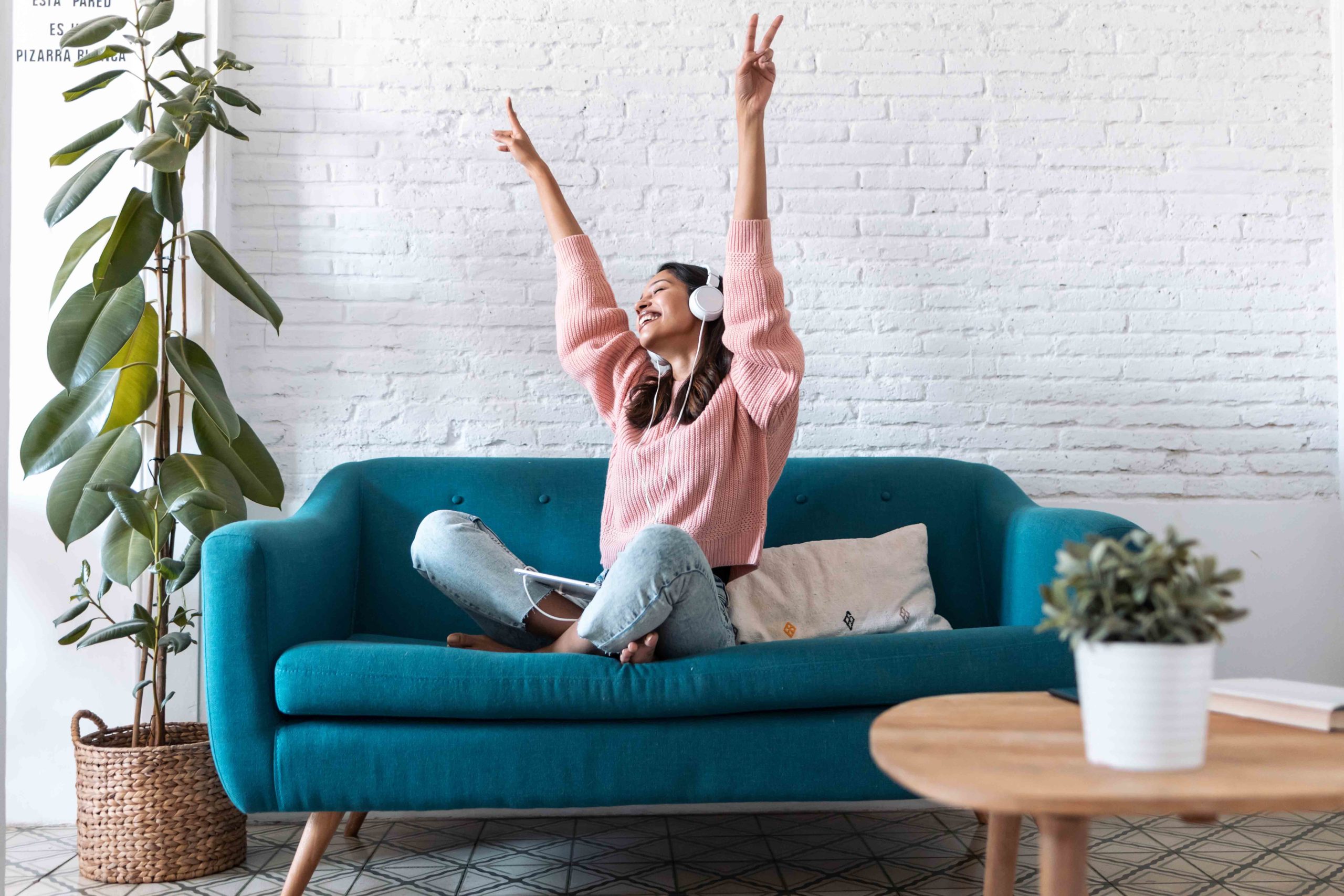 A joyful woman wearing headphones is sitting on a teal sofa with her hands raised in the air, seemingly enjoying her favorite music in a bright, cozy living room.