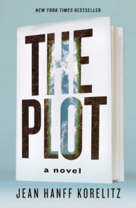 A hardcover book titled "the plot" by jean hanff korelitz stands against a pastel blue background. the cover features a nature-inspired design with elements of forest and sky visible through the cut-out letters of the title.