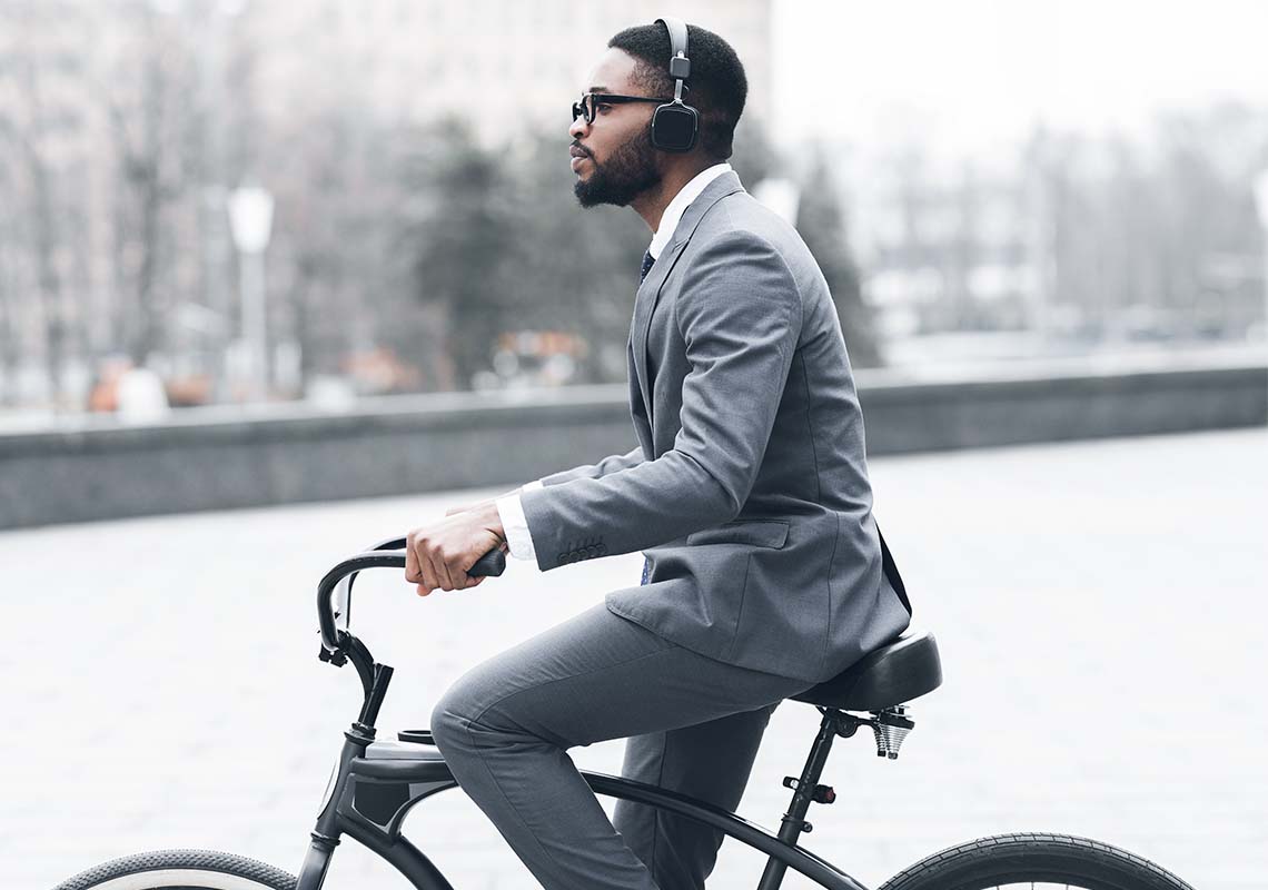 A business professional rides a bicycle while wearing headphones and a stylish gray suit, showcasing a modern blend of fitness-conscious commuting and sharp urban fashion.