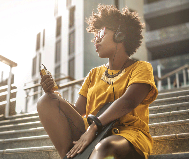 A young person sitting on steps, enjoying music on headphones, and holding a mobile device in a sunny urban setting.