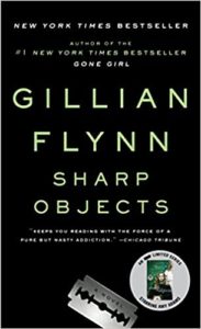 A book cover of gillian flynn's novel "sharp objects" featuring the title and author's name in large, bold prints, with critical acclaim quotes and an award sticker.
