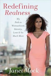An individual in a stylish pink dress stands confidently with a cityscape in the background, on the cover of a book titled "redefining realness: my path to womanhood, identity, love & so much more" by janet mock.
