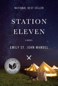 A starlit night sky forms the background for the book cover of "station eleven" by emily st. john mandel, prominently featuring a lit tent and accolades such as national bestseller and national book award finalist.