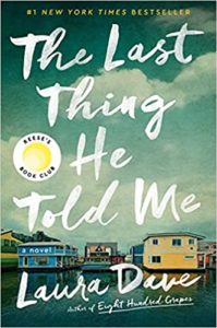 A book cover for the novel "the last thing he told me" by laura dave, featuring a waterfront scene with a house and boat docked by the shore, set against a backdrop of fading handwritten text in the sky.