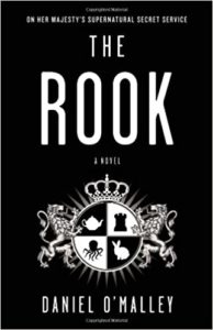 A black and white book cover for "the rook" by daniel o'malley, featuring a stylized coat of arms with a chess rook piece, a knight, a crown, and shadowy figures, set against a dark background with the title and author's name prominently displayed.