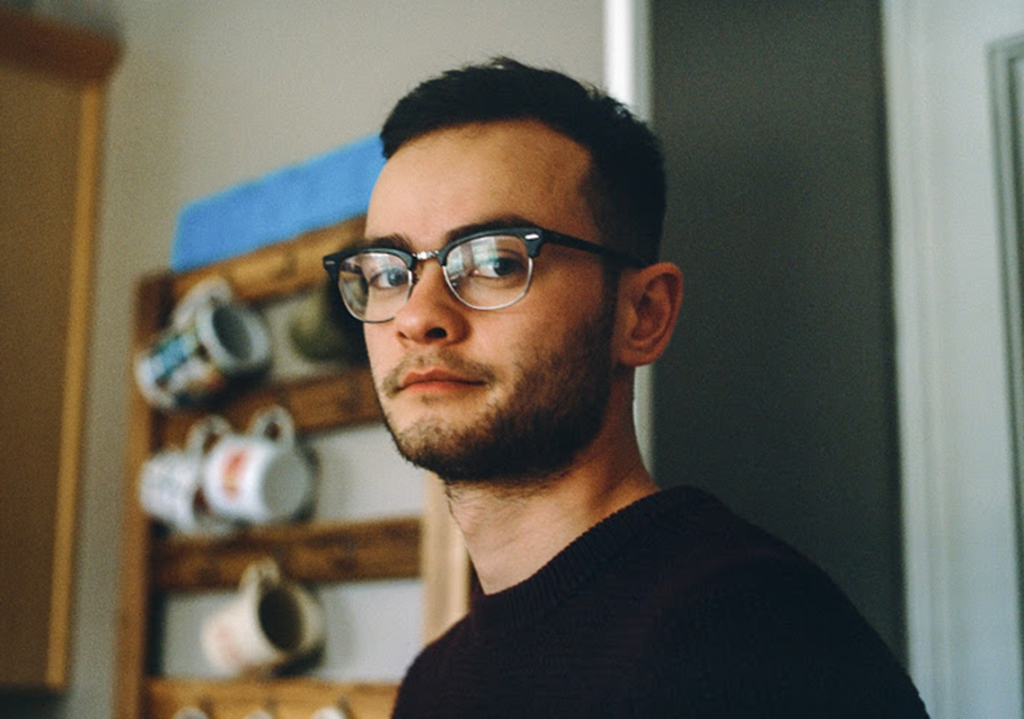 A pensive young man with glasses, standing indoors and gazing off into the distance, with a blurred background featuring a wall-mounted shelf holding mugs.