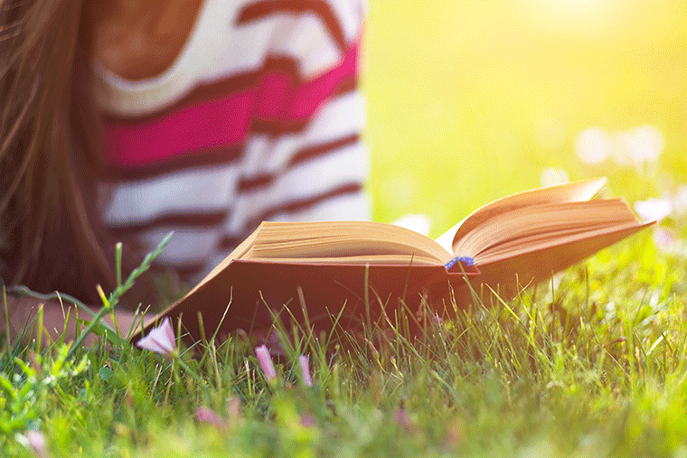 A person lounges on the grass, deeply absorbed in an open book on a sunny day, surrounded by the warmth of sunlight and the serenity of nature.