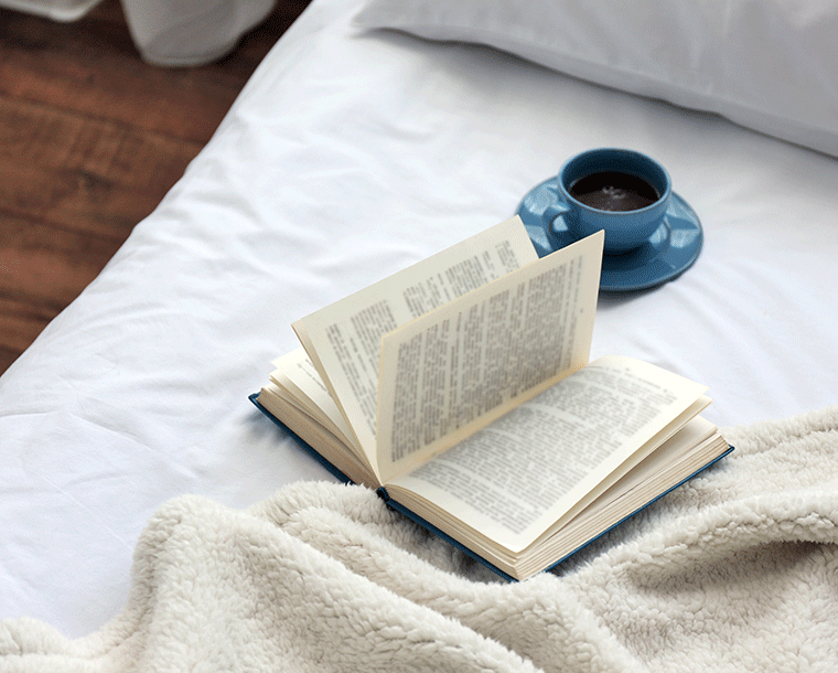 A cozy reading nook with an open book resting on a soft blanket, accompanied by a cup of hot beverage on a bedside table, inviting relaxation and leisure.