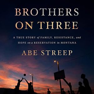 A silhouette of basketball players at sunset with the title "brothers on three" by abe streep, highlighting a story of family, resistance, and hope on a reservation in montana.