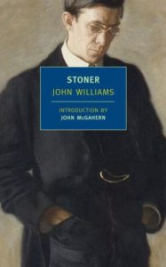 A man in a formal dark suit with a pensive expression holds a book titled "stoner" by john williams, with an introduction by john mcgahern.