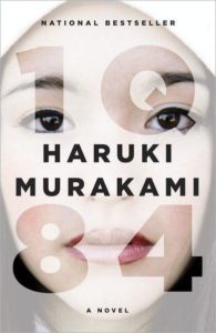 A conceptual book cover design for haruki murakami's novel "1q84," featuring a close-up of a woman's face partitioned by the title text in large bold numbers and letters with the author's name beneath, all overlaying the image in a manner that aligns elements of the face with the spaces in the text.