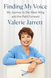 A woman with a confident smile, wearing a blue cardigan, sits relaxed with her arms resting on the back of a chair, posing for the cover of her book titled "finding my voice: my journey to the west wing and the path forward" by valerie jarrett.