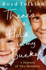 A book cover titled "there's a hole in my bucket" by royd tolkien, featuring a close-up photo of two smiling young boys, one slightly older than the other, with the older boy wearing a red ensemble and the younger in a blue outfit, symbolizing the bond and journey of two brothers.