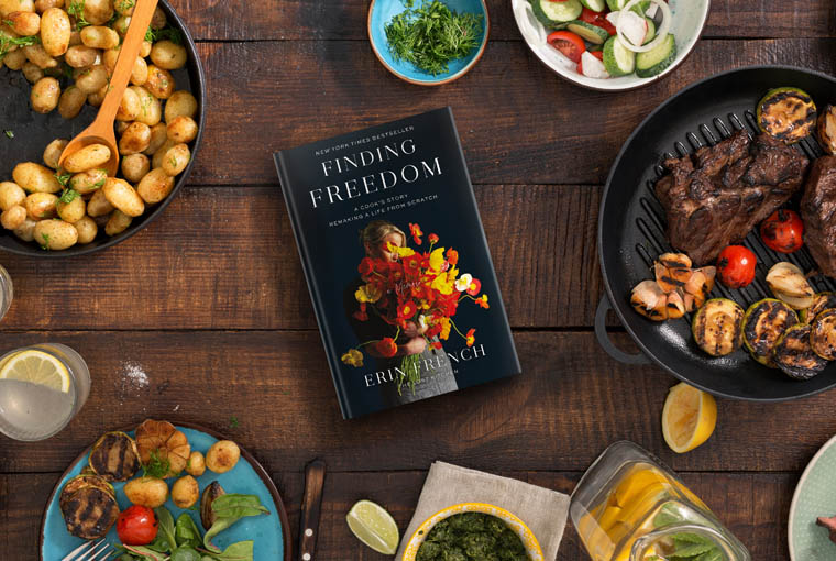 A well-arranged dining table with a variety of dishes including grilled steak, shrimp, and potatoes, complemented by fresh vegetables and herbs, featuring a book titled "finding freedom" by erin french as the centerpiece amongst the meal.