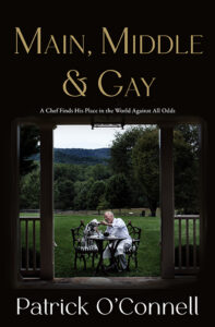 Chef sits at an outdoor table with a dog, mountains and trees in the background. Book title above.