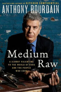 A thoughtful man in a suit with crossed arms leaning on a kitchen counter, featured on the cover of the book "medium raw" by anthony bourdain.