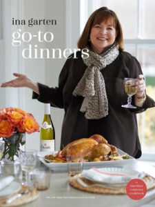 A smiling person gesturing towards a dining table set with a roasted chicken, a bottle of wine, and a bouquet of flowers, with a book cover stating "go-to dinners" by a new york times bestselling author.