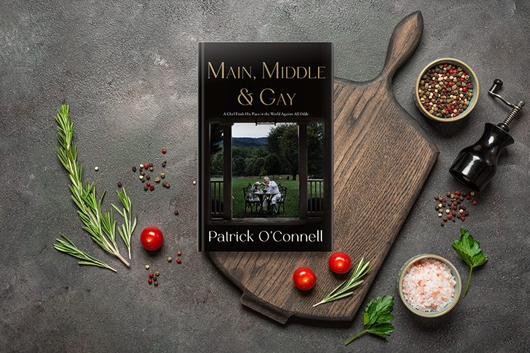 A cookbook on a wooden board, surrounded by herbs, cherry tomatoes, salt, and pepper mills.