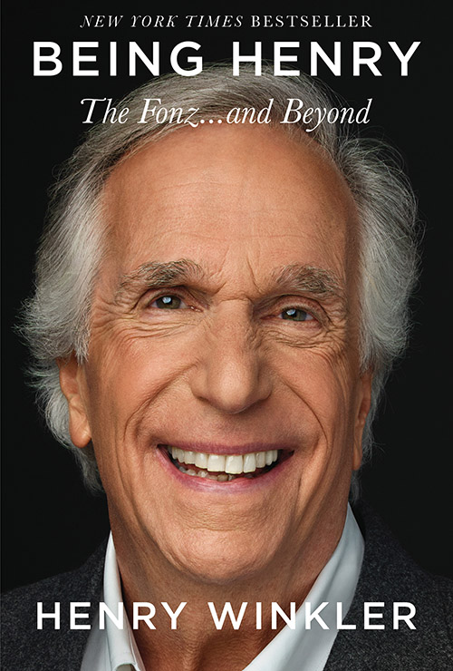 A portrait of a cheerful older man with a broad smile, captioned with the title of his book "being henry - the fonz...and beyond," suggesting an autobiography by the man pictured, who has a notable presence and charisma.