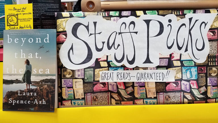 A colorful display of books recommended by staff at a bookstore, featuring a prominent sign that reads "staff picks - great reads - guaranteed!!" with a showcased book titled "beyond that, the sea" by laura spence-ash.