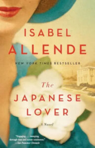 A close-up of a book cover titled "the japanese lover" by isabel allende, featuring a partial profile of a woman's face with red lipstick, positioned behind soft white flowers, with a small image of a classic building in the background, accompanied by quotes praising the novel.