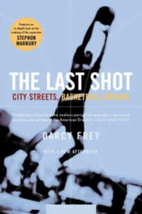 A book cover for "the last shot: city streets, basketball dreams" by darcy frey, featuring an out-of-focus background of a basketball hoop with the silhouette of a basketball player in mid-jump, capturing the essence of urban basketball aspirations.