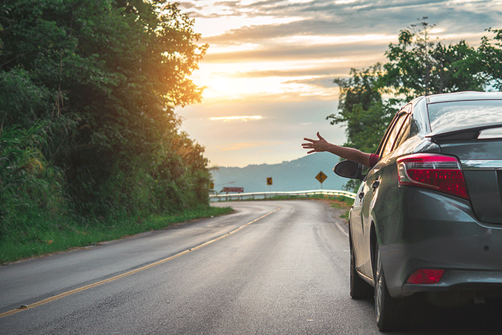 A serene road trip moment: a passenger leans out of a car window, feeling the breeze, on a winding road at sunset.